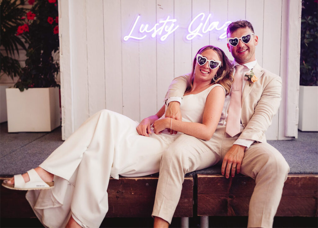 Wedding Couple sat down on stage in front of a Lusty Glaze neon sign. The bride and groom are wearing identical sunglasses that have their initials on the left lens and wedding date on the right lens. Photo Credit Albion Row.